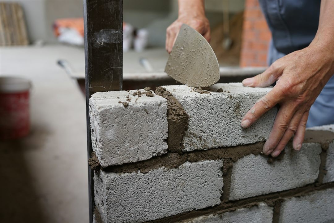 A bricklayer buttering mortar with a trowel and bedding a concrete block onto a wall against a level line