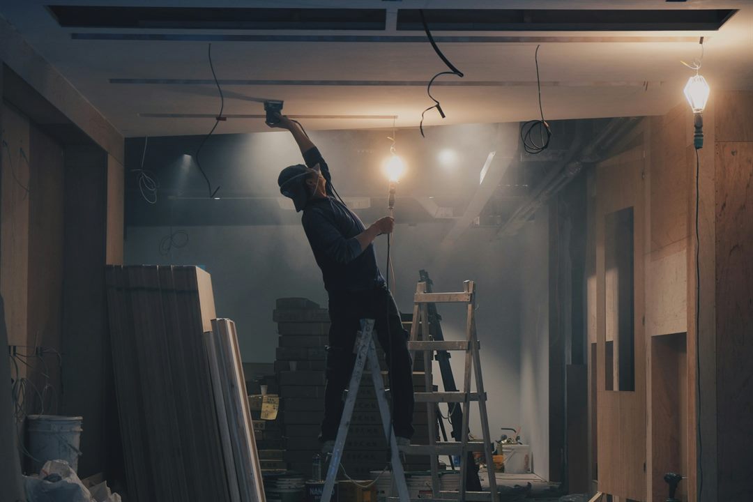 Electrician on a stepladder fixing lighting to the ceiling of a strip-out fit-out, temporary festoon lights hanging