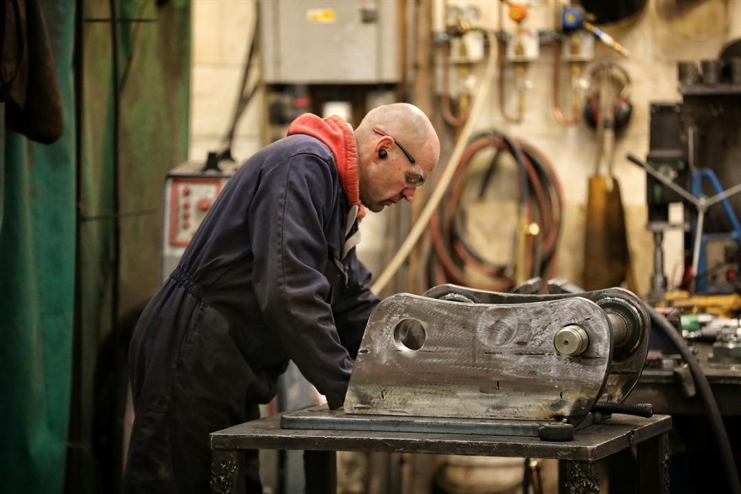 Fabricator in overalls and safety glasses working on a machined steel bracket at the bench